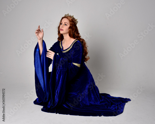 Full length portrait of  girl wearing long blue velvet gown with golden crown. sitting pose, isolated against a studio background.