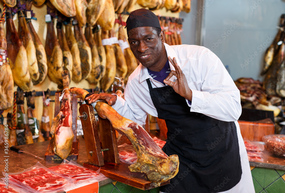 Happy African American seller working in meat shop, recommending delicious iberian ham..