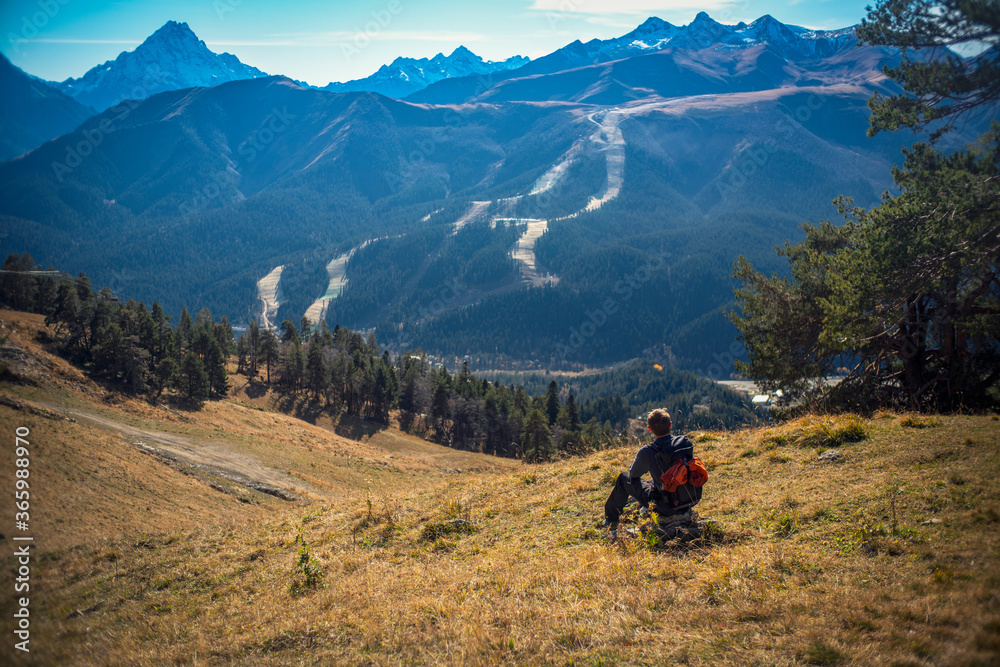 A tourist sits in a clearing and looks into the distance. Nearby are large conifers, below the trees there is dry grass and fallen cones, mountains are visible in the distance.