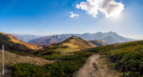 Panoramic photo of a mountain valley, people are walking down between the mountains, clouds are floating in the sky and the mountains are covered with greenery, there is warm sunny weather.
