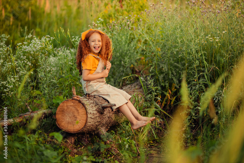 A cheerful, perky red-haired girl sits on a log on a summer walk in the forest.