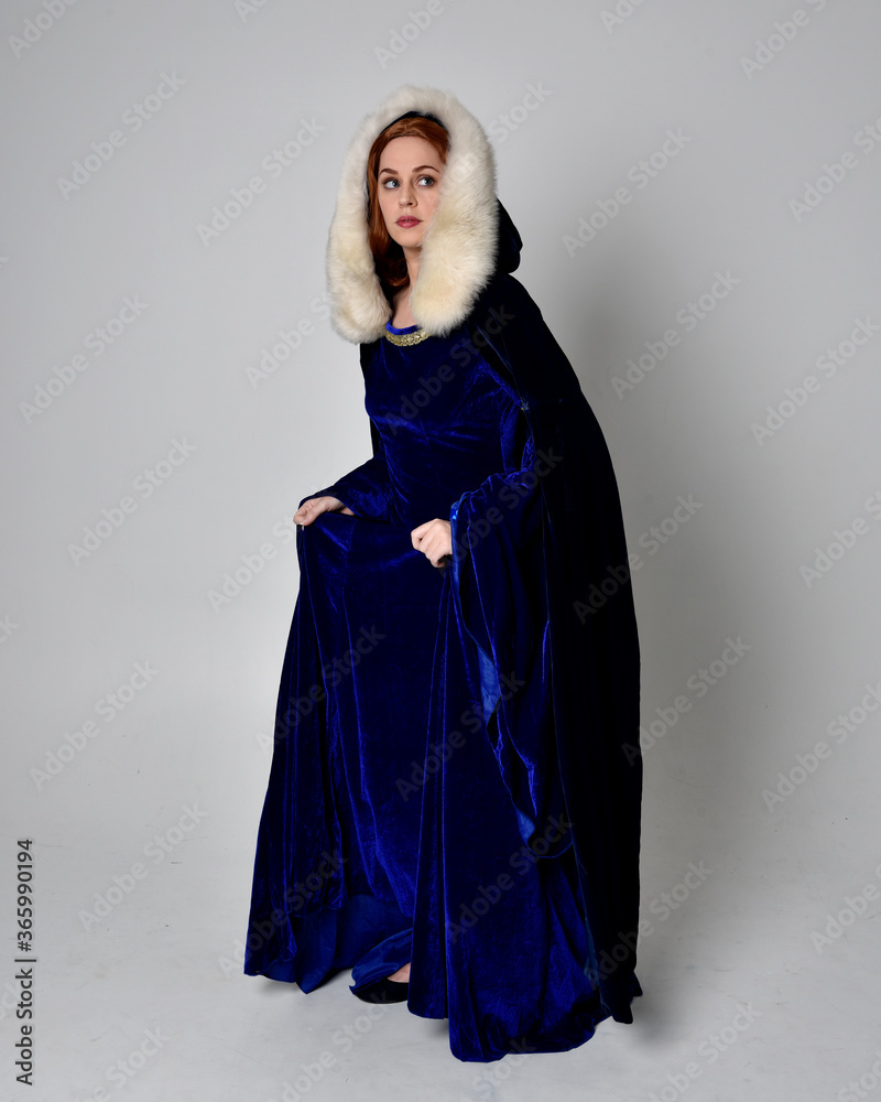 Full length portrait of  girl wearing long blue velvet gown with golden crown. sitting pose, isolated against a studio background.