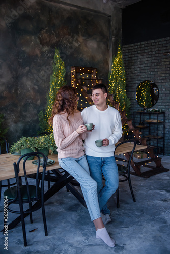 A loving couple in warm sweaters hold two turquoise ceramic cups in their hands, drink hot drinks in winter, against the background of Christmas lights. The atmosphere of winter holidays at home