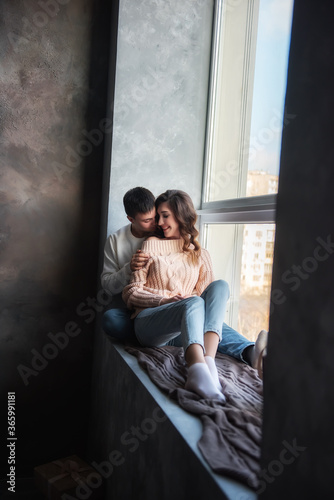 Happy couple in love in winter sweaters and jeans, sit on a wide windowsill by the window, hug. A young man kisses a beautiful curly brown-haired girl on the cheek. Date by the fireplace in cozy home