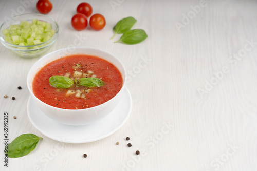 White bowl of gazpacho soup made of red pureed tomatoes decorated with basil leaves standing on light wooden table at kitchen. Image with copy space. horizontal orientation