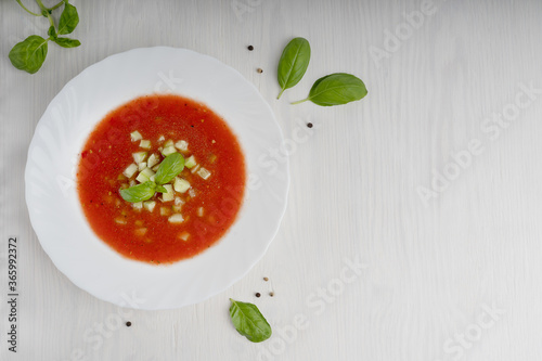 White plate with red gazpacho soup made of pureed tomatoes and other vegetables standing on light wooden table at kitchen surrounded by basil leaves. Image with copy space, horizontal orientation