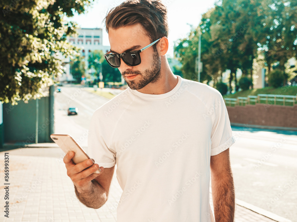 Handsome smiling stylish hipster lambersexual model.Man dressed in white T-shirt.Fashion male posing in the street. Holding, scrolling and texting in his cellphone. Writes sms on smartphone Handsome smiling stylish hipster lambersexual model.Man dressed in white T-shirt.Fashion male posing in the street. Holding, scrolling and texting in his cellphone. Writes sms on smartphone
