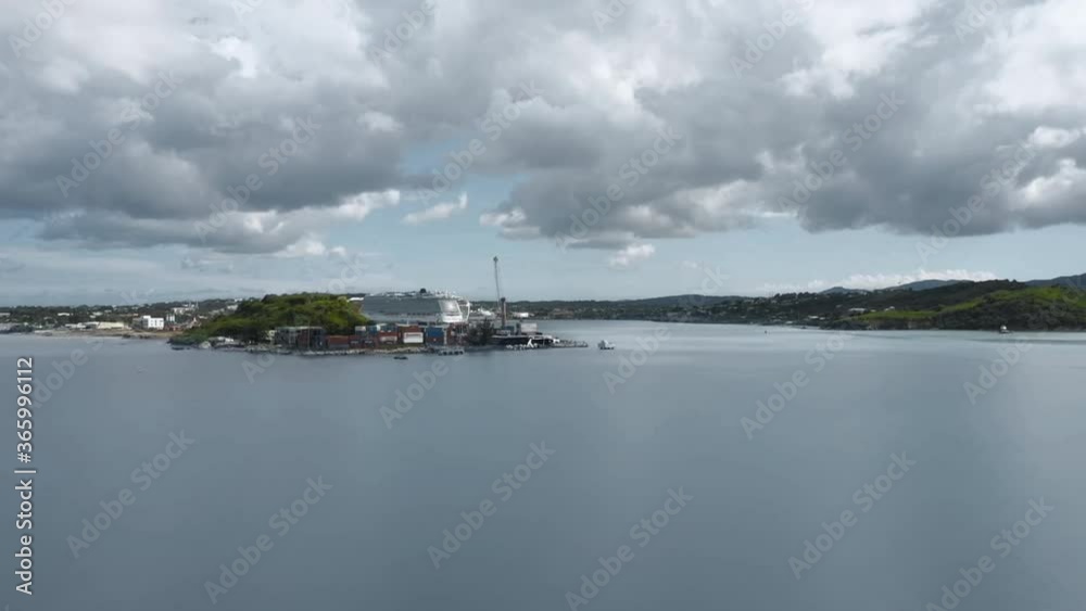 Drone is approaching a bay with a cruise ship. Hilly island. A lot of clouds. Aerial shot.