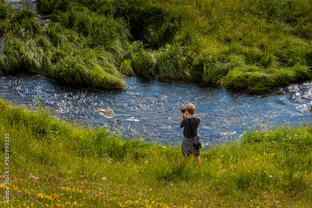 young man taking photos next to a river in the countryside, in summer .with wild flowers in forground.