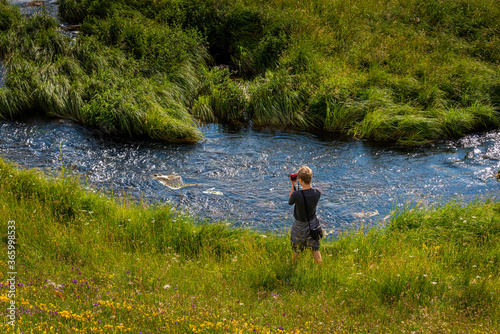 young man taking photos next to a river in the countryside, in summer .with wild flowers in forground.