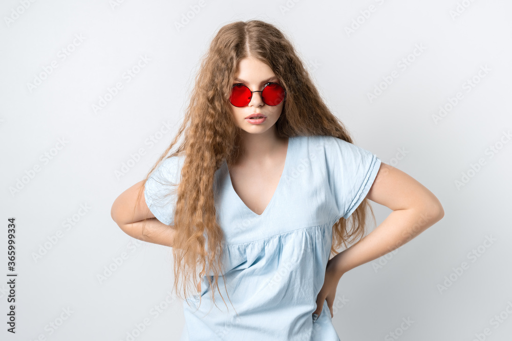 Portrait of charismatic modern girl with curly long hair in casual light clothes and wears round red spectacles posing isolated on white wall background. People lifestyle concept Portrait of charismatic modern girl with curly long hair in casual light clothes and wears round red spectacles posing isolated on white wall background. People lifestyle concept
