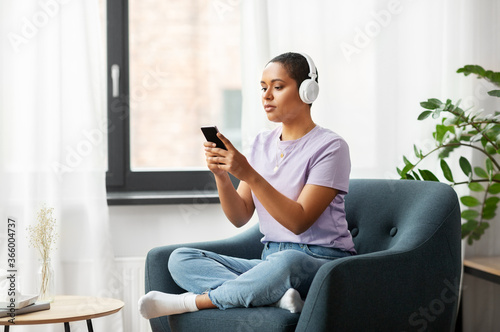 people, technology and leisure concept - young african american woman in glasses with smartphone and headphones sitting in chair and listening to music at home
