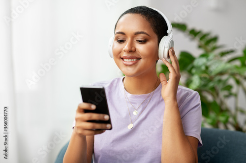 people, technology and leisure concept - happy smiling young african american woman in glasses with smartphone and headphones sitting in chair and listening to music at home