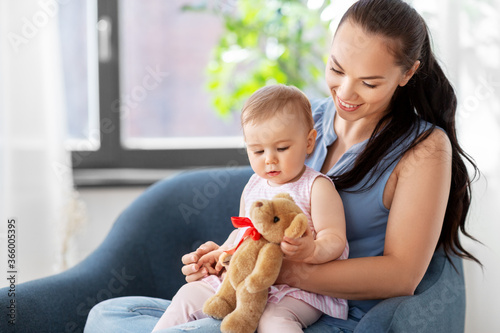 family, child and motherhood concept - portrait of happy smiling mother and little baby daughter with teddy bear toy sitting in chair at home