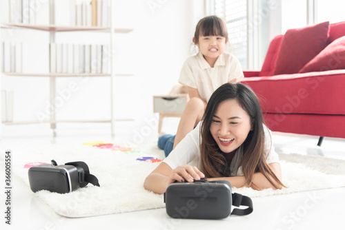 Happiness young Asian mother and daughter lie on floor of house, the child riding on the back of the mother, they play VR game for entertain , family joyful together in the house