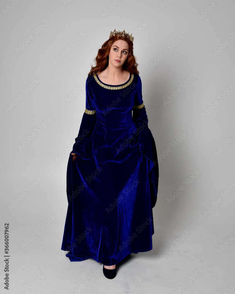 Full length portrait of  girl wearing long blue velvet gown with golden crown. standing pose with back to the camera, isolated against a studio background.