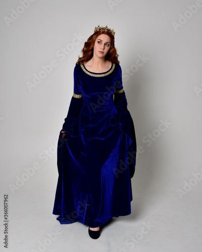 Full length portrait of  girl wearing long blue velvet gown with golden crown. standing pose with back to the camera, isolated against a studio background.