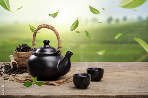 Cup of hot tea with teapot, flying green tea leaves in the air and dried herbs on the wooden table in plantations background, Organic product from the nature for healthy with traditional