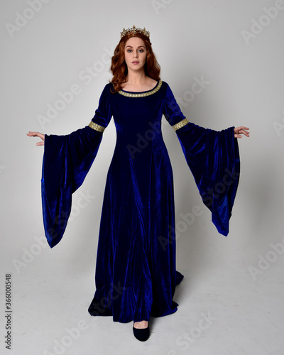 Full length portrait of  girl wearing long blue velvet gown with golden crown. standing pose with back to the camera, isolated against a studio background.