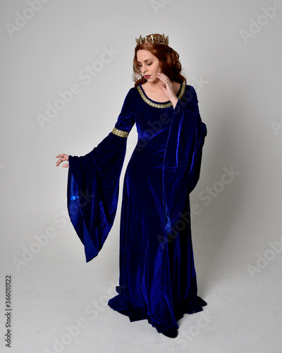 Full length portrait of  girl wearing long blue velvet gown with golden crown. standing pose with back to the camera, isolated against a studio background.