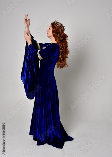 Full length portrait of girl wearing long blue velvet gown with golden crown. standing pose with back to the camera, isolated against a studio background.