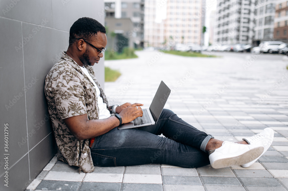 Distant learning. Handsome stylish African American student in sunglasses dressed in a casual wear sits outdoors and learns remotely using his laptop