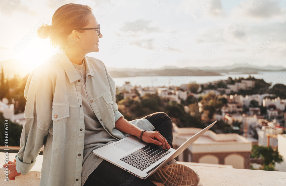 Beautiful curly young girl with glasses working on a laptop with a city view at sunset. Modern technologies, urban lifestyle. Freelance and online education