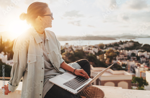 Obraz na płótnie Beautiful curly young girl with glasses working on a laptop with a city view at sunset. Modern technologies, urban lifestyle. Freelance and online education w salonie