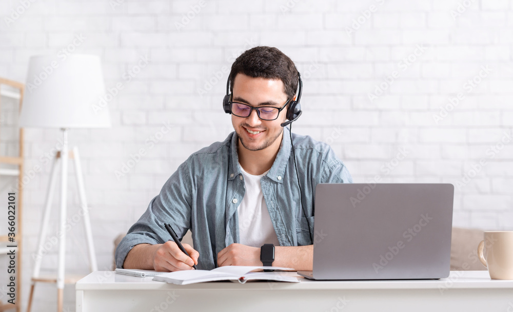 Education remotely and independently. Smiling guy with headphones makes notes in notebook with laptop on desktop Education remotely and independently. Smiling guy with headphones makes notes in notebook with laptop on desktop