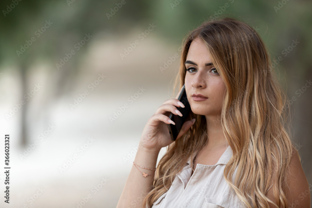 Portrait of young blonde italian woman talking at the phone inside the park whit shallow depth of field with blurred tree in the background