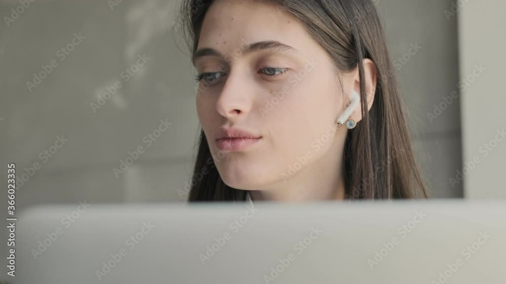 Young concentrated business woman indoors at home using laptop computer while listening music with wireless earphones