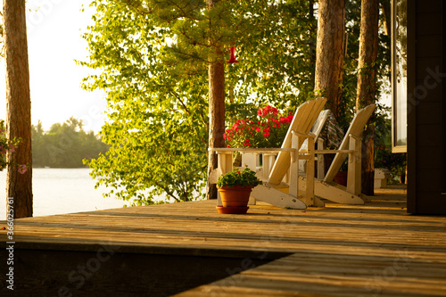 White Adirondack chairs sits on a cottage wooden deck at sunset looking at the lake. Flower pots are on the deck.