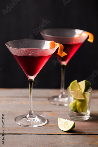 Closeup 2 glasses of cosmopolitan cocktail decorated with orange served with lime wedges.  Dark grey worktop with dark background