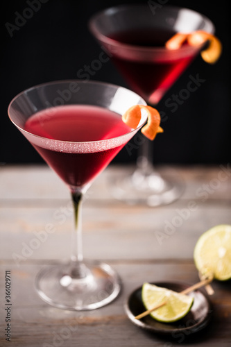 Closeup 2 glasses of cosmopolitan cocktail decorated with orange served with lime wedges.  Dark grey worktop with dark background