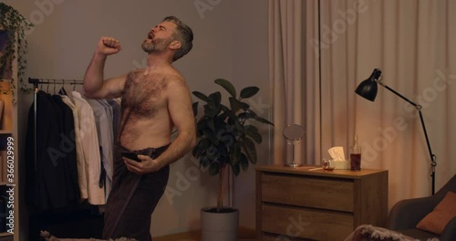 Handsome guy in 40s pretending playing guitar and singing at home. Mature cheerful man in bath towel having fun while looking at smartphone and typing. Concept of leisure. photo