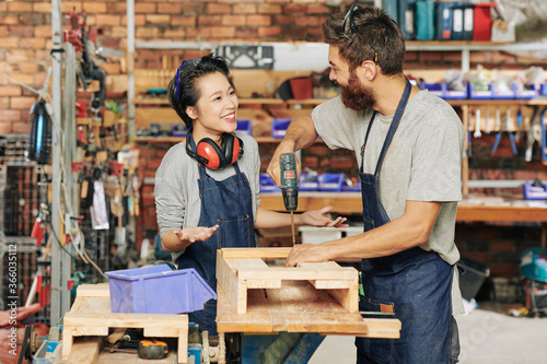 Smiling female carpenter talking to her coworker drilling screws when making wooden drawer