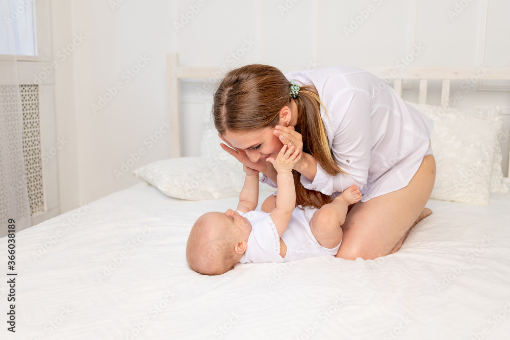 mother sits with baby 6 months on a white bed at home and plays with it, place for text mother sits with baby 6 months on a white bed at home and plays with it, place for text