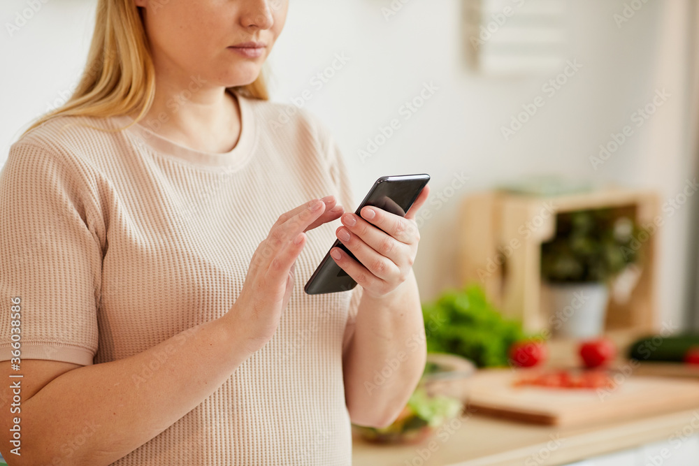 Cropped portrait of adult woman using smartphone and typing text message while standing in cozy kitchen interior, copy space Cropped portrait of adult woman using smartphone and typing text message while standing in cozy kitchen interior, copy space