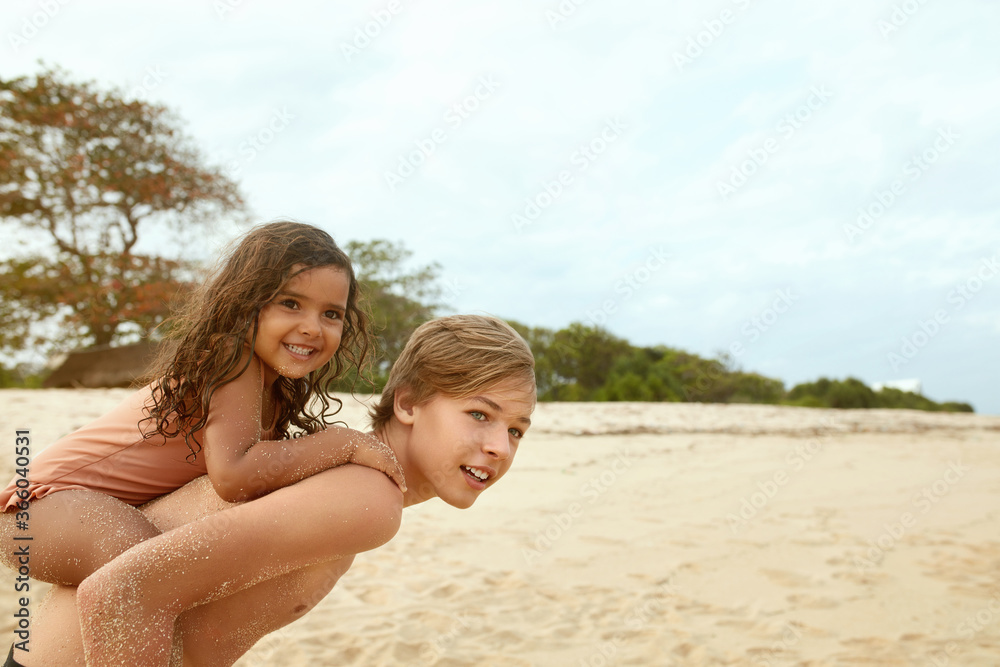 Boy And Girl On Beach. Tanned Sister On Brother's Back Spending Time On Coast. Positive Siblings Covered With Sand And Enjoying Summer Vacation. Happy Childhood And Active Recreation. Boy And Girl On Beach. Tanned Sister On Brother's Back Spending Time On Coast. Positive Siblings Covered With Sand And Enjoying Summer Vacation. Happy Childhood And Active Recreation.