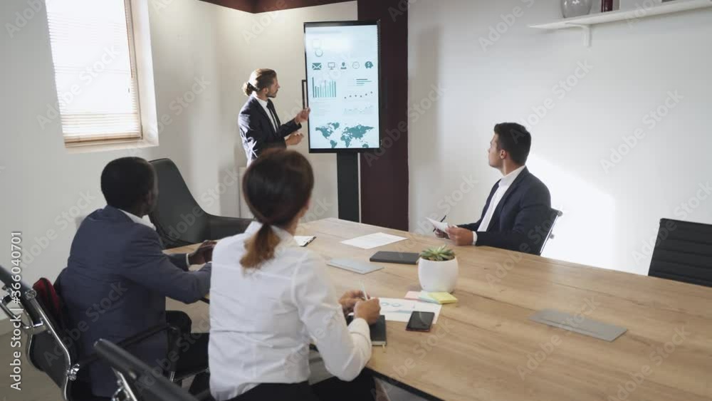 Internationl business meeting, young businessman speak at a meeting and shows financial infographic in the conference room, man in suit communicates with colleagues, infographics on vertical screen. Internationl business meeting, young businessman speak at a meeting and shows financial infographic in the conference room, man in suit communicates with colleagues, infographics on vertical screen.