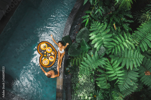 Travel happy couple in love eating floating breakfast in jungle swimming pool. Awakening in morning. Black rattan tray in heart shape, Valentines day or honeymoon surprise, view from above. photo