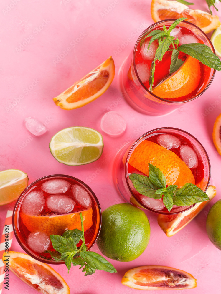 Naklejka premium Fresh iced red cocktail in pink glasses with blood orange and lime on pink background garnished with mint leaves. Closeup