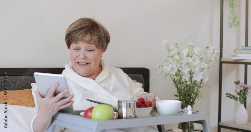 Cheerful mature lady in rob having video call during breakfast in bed. Good looking woman pensioner communication on tablet using frontal camera while sitting with trey full of food. Cheerful mature lady in rob having video call during breakfast in bed. Good looking woman pensioner communication on tablet using frontal camera while sitting with trey full of food.