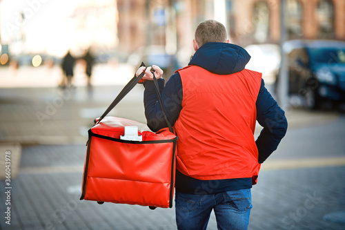 Man delivering food to costumers, walking with red thermal bag on city street, grocery delivery. Boy deliver food online orders to customer. Courier carries red bag with fresh food. Delivery service