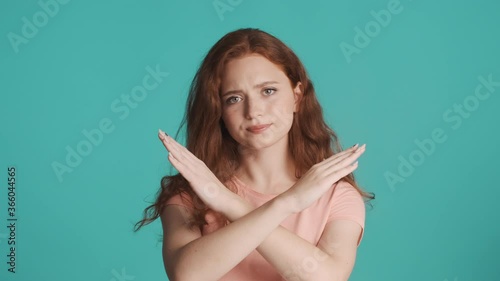Serious redhead girl confidently showing no gesture with crossed hands over blue background. No way expression