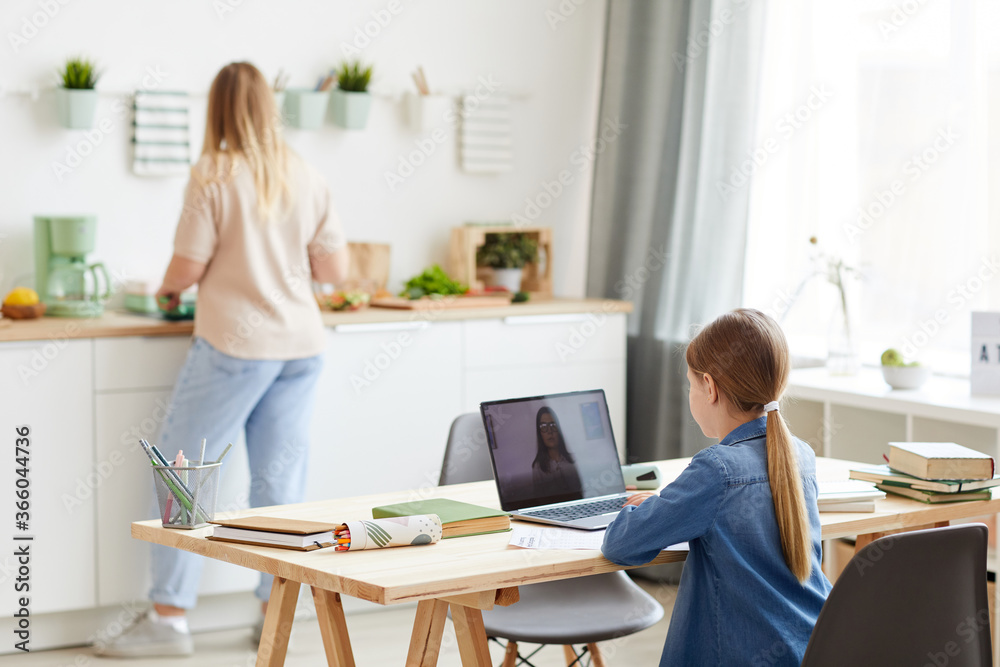 Back view portrait of little girl using laptop during online class with tutor or teacher while sitting at desk in cozy kitchen interior with mother in background, copy space Back view portrait of little girl using laptop during online class with tutor or teacher while sitting at desk in cozy kitchen interior with mother in background, copy space