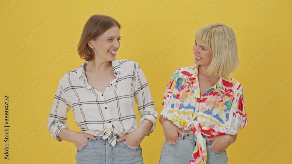 Two happy pretty women having fun together with arms in pockets over yellow background Two happy pretty women having fun together with arms in pockets over yellow background