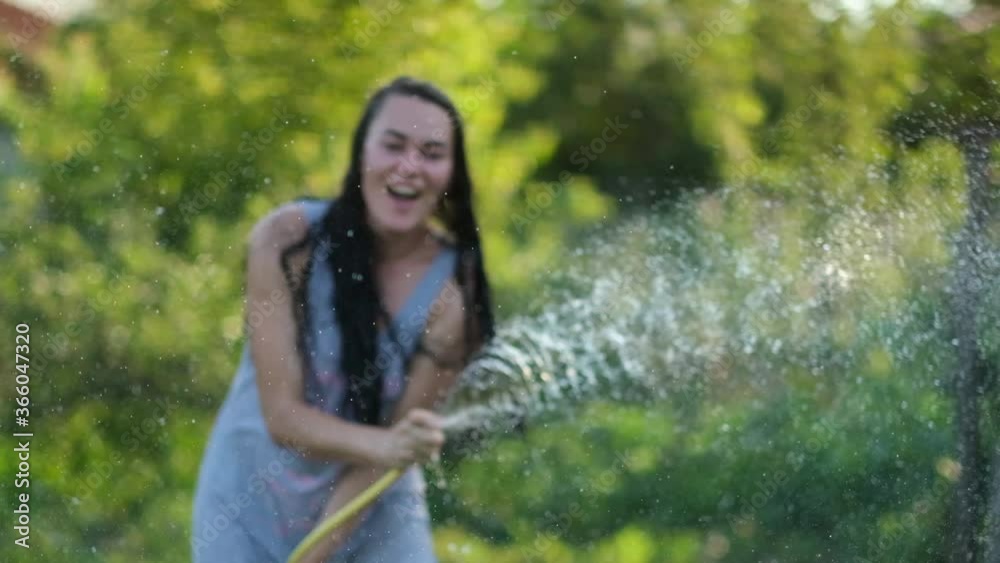 Happy sexy woman girl spraying with water on a hot summer day holding a hose pipe above her head in a beam of warm light from the sun near garden trees and plants. Slow motion Happy sexy woman girl spraying with water on a hot summer day holding a hose pipe above her head in a beam of warm light from the sun near garden trees and plants. Slow motion