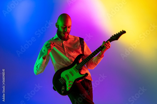 Tensioned. Young caucasian inspired and expressive musician, guitarist performing on multicolored background in neon. Concept of music, hobby, festival, art. Joyful artist, colorful, bright portrait.