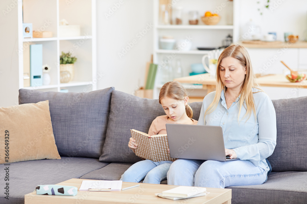 Portrait of adult working mother using laptop while sitting on couch at home with cute daughter reading book beside her, copy space Portrait of adult working mother using laptop while sitting on couch at home with cute daughter reading book beside her, copy space